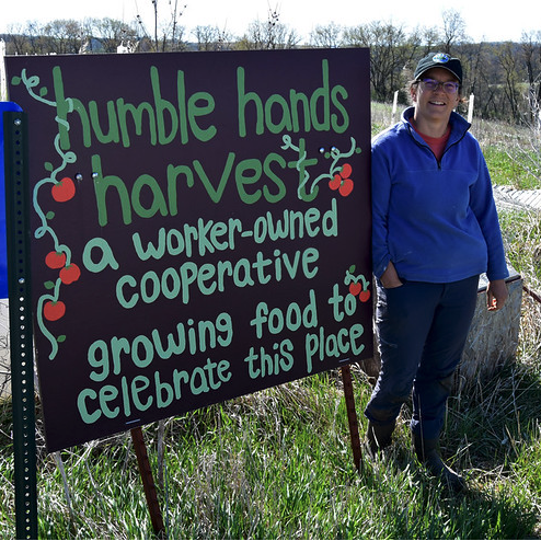Hannah Breckbill standing in front of sign reading "humble hands harvest - a worker owned cooperative growing food to celebrate this place."